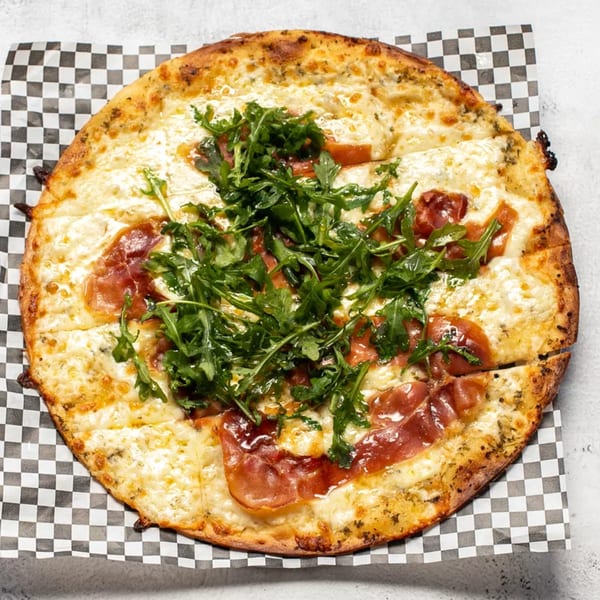 A whole pizza with white cheese, prosciutto, and arugula, on checkered paper.