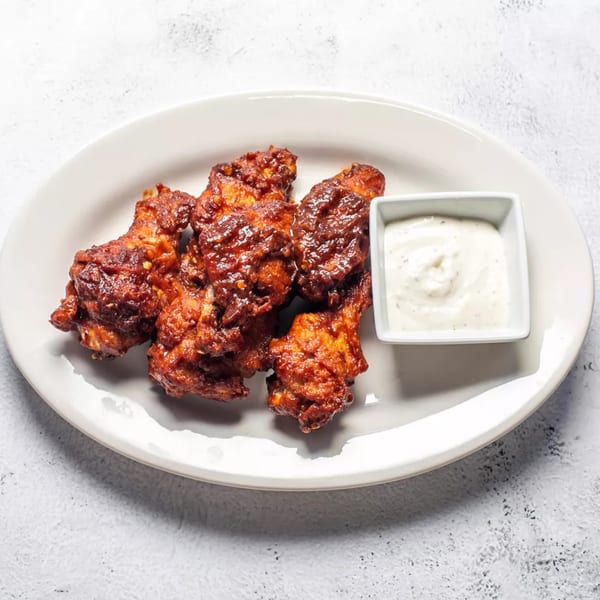 A white oval plate holds five dark red, saucy chicken wings and a small square dish of ranch dressing.